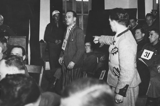Un martor al acuzării îl indică pe acuzatul Emil Erwin Mahl în timpul procesului pentru crime de război de la Dachau. Mahl a fost condamnat pentru crimele de război pe care le-a comis în calitate de kapo, inclusiv pentru că a ascultat de ofițerii SS și a legat ștreangurile la gâtul prizonierilor